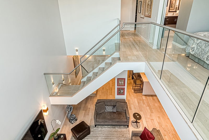 Elegant open-concept living space with a floating staircase featuring glass railings, modern lighting, and a loft-style upper level, designed by Sayers Contracting.