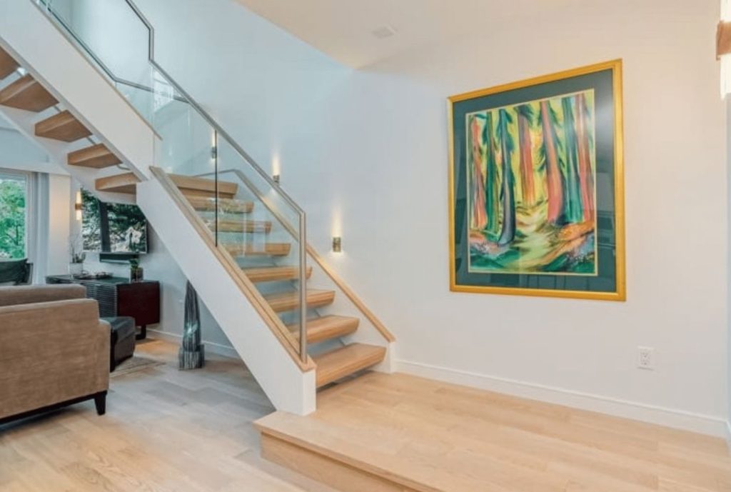 Modern home interior with floating wooden staircase, glass railing, light hardwood floors, and contemporary wall art.