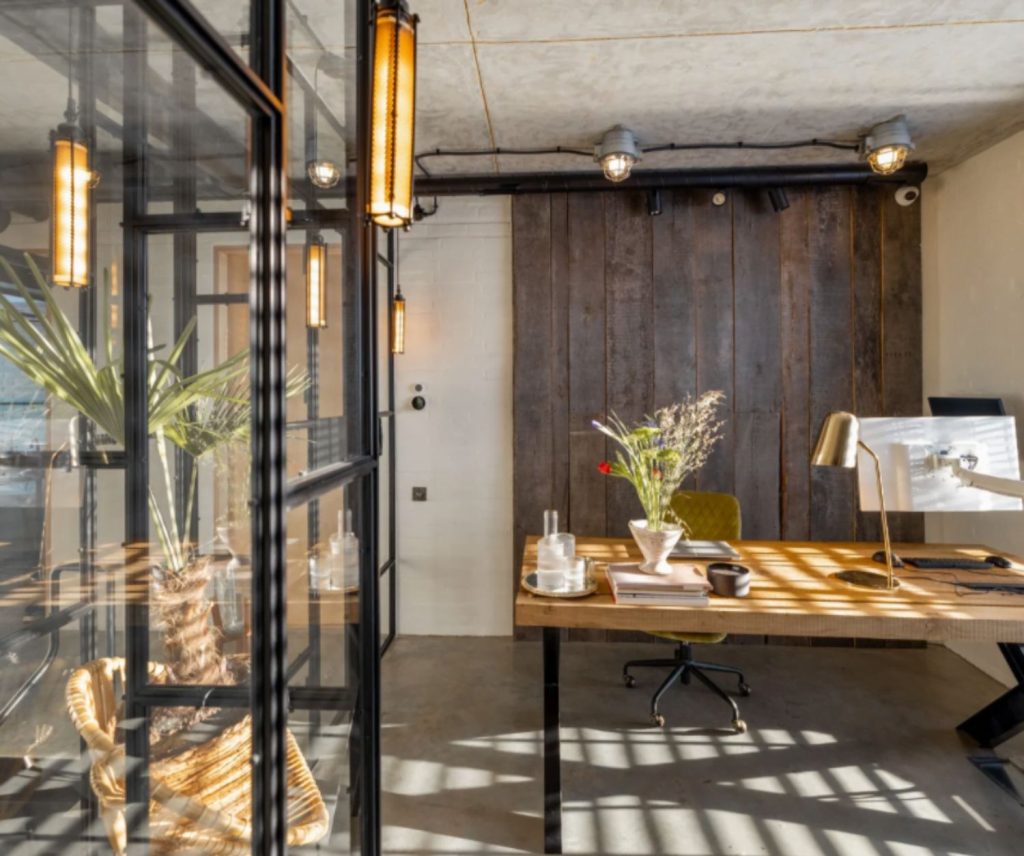 Modern industrial-style home office with a wooden desk, metal-framed glass partitions, rustic wood paneling, and natural lighting.
