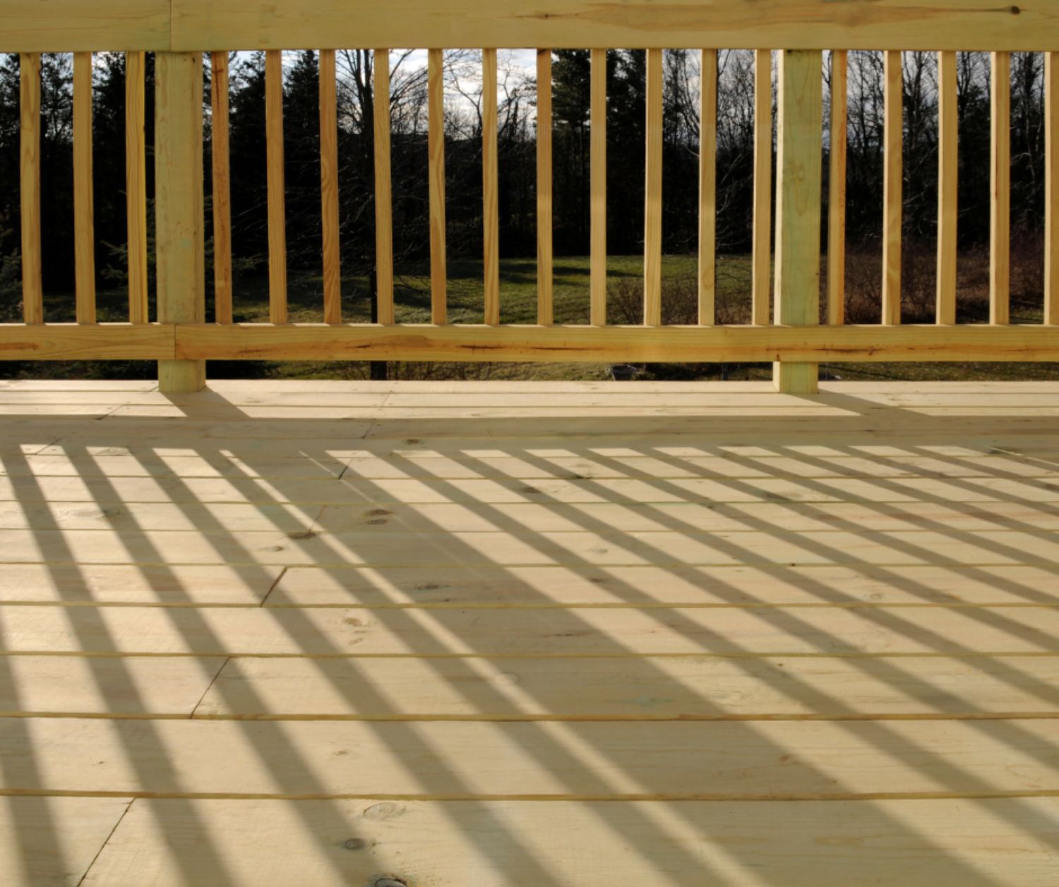 Newly built wooden deck with railing, casting long shadows in natural sunlight, overlooking a scenic outdoor landscape.